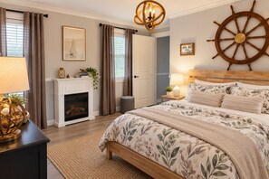 Une chambre à coucher avec une cheminée, un lit recouvert d’une literie à motifs floraux, une décoration en forme de barre de gouvernail de bateau et un lustre.