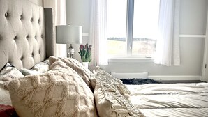 Ein Schlafzimmer mit einer gesteppten Kopfteil, einer Nachttischlampe, einer Vase mit Blumen und Blick ins Freie durch ein Fenster.