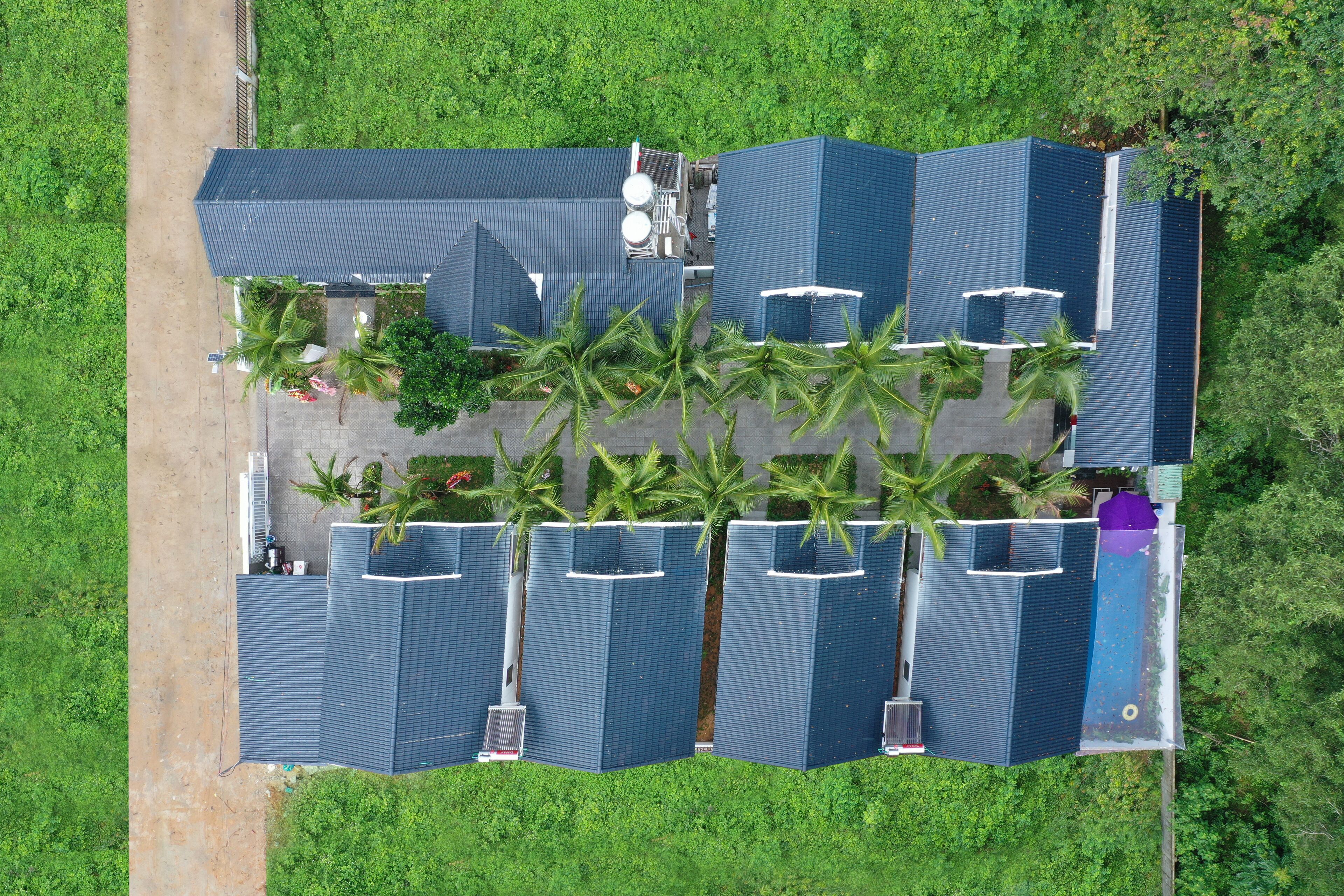 An aerial view of a hotel with multiple blue-roofed buildings, arranged in a U-shape, surrounded by lush greenery and palm trees.