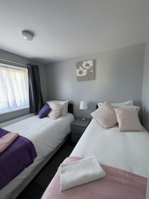 A hotel room with two beds, a nightstand, a lamp, and a window with curtains. - Mona’s AirB&B in quiet cul-de-sac. 3 bedrooms, 4 beds plus  sofabed. Sleeps 6.  (Dagenham)