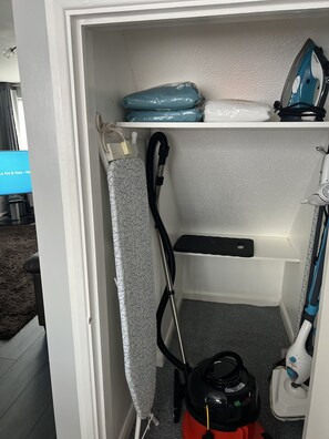 A storage closet with shelves holding cleaning supplies, an iron, and a vacuum cleaner.