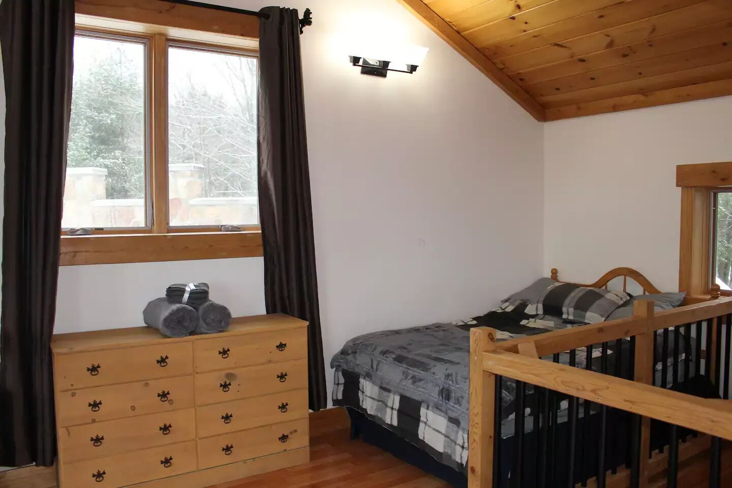 A bedroom with a wooden dresser, a bed with a headboard, a window with curtains, and a wall-mounted light fixture.
