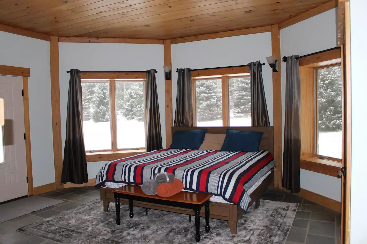 Um quarto acolhedor com teto de madeira, cama com roupa de cama listrada, duas janelas com cortinas e um tapete com padrão de pedra.