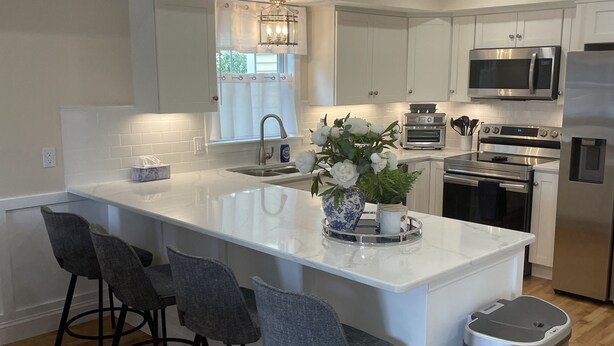 A modern kitchen with white cabinetry, stainless steel appliances, a central island with bar stools, and a trash can.