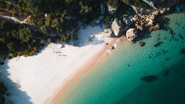 Vue aérienne d’une plage aux eaux turquoise et limpides, au sable blanc et aux formations rocheuses.