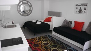 Une chambre d’hôtel moderne avec une kitchenette blanche, deux lits avec une literie noire et rouge, un tapis à motifs et un miroir fixé au mur.