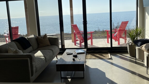 Ein modernes Wohnzimmer mit einem großen Schiebeglasfenster, das auf eine Terrasse mit roten Stühlen mit Blick aufs Meer führt.