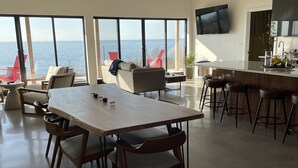Eine moderne Küche mit einer großen Kochinsel, einem Essbereich mit Holztisch und -stühlen sowie großen Fenstern mit Meerblick.