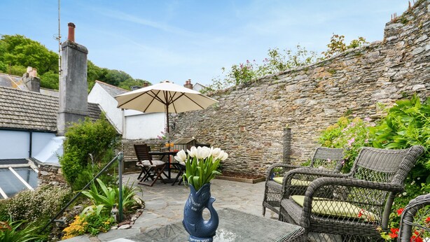 A patio area with a stone wall, wicker furniture, a table with chairs, and a vase of white flowers. - Wedgwood Cottage - Sleeps 6 guests  in 3 bedrooms (Cawsand)