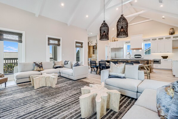 A spacious living room with a large sectional sofa, a coffee table, and a dining area with a kitchen in the background. - 3-story ocean-view home with pool, grill, golf, gym, pool table, & washer/dryer (Port Aransas)