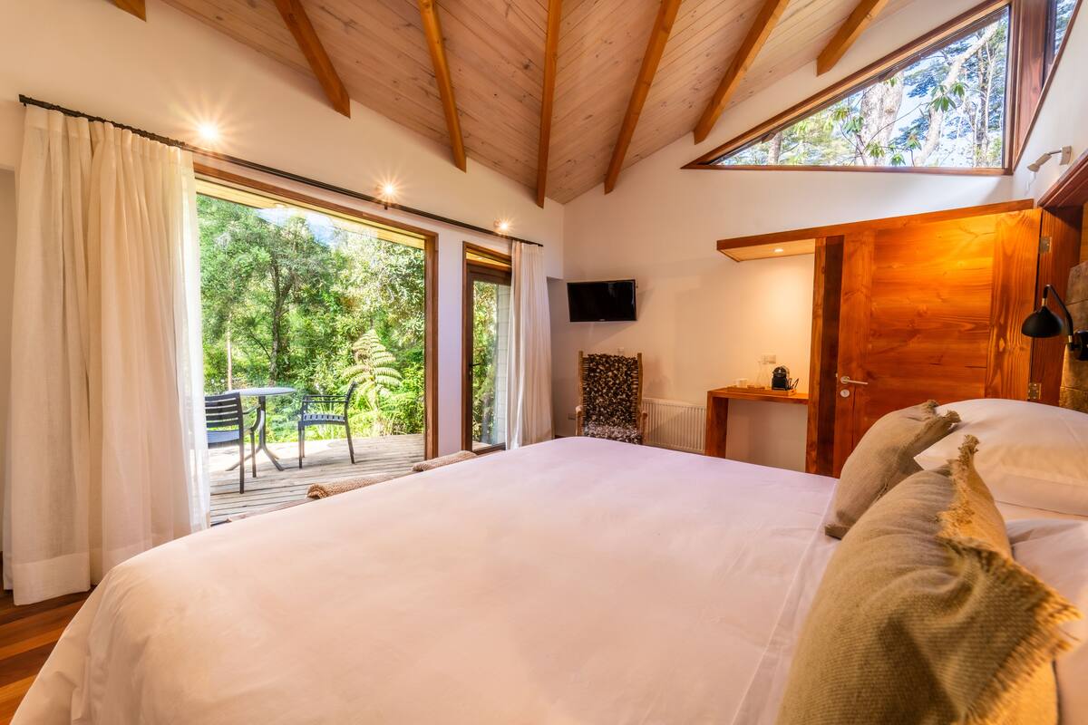 A bedroom with a large bed, wooden ceiling beams, a TV, and a view of greenery through the windows.