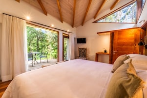 Un dormitorio con una cama grande, vigas de madera en el techo, televisión y vistas a la vegetación por las ventanas.