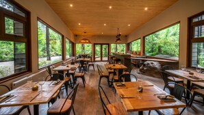 A restaurant with wooden tables and chairs, large windows offering views of trees, and a bar area with bottles. - rakau lodge (Pucón)