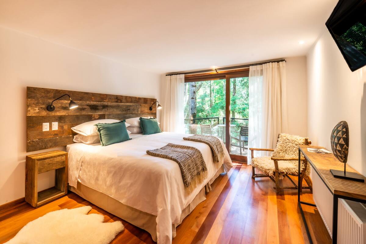 A bedroom with a large bed, a wooden headboard, a desk with a chair, and a view of greenery through the window.