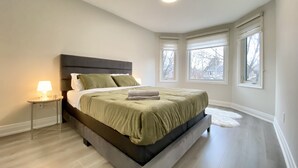 A modern bedroom with a large bed, a nightstand, and a window with a view of trees. - Waterfront Paradise Estate (Aurora)