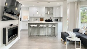 A modern kitchen with white cabinets, a central island with bar stools, a flat-screen TV mounted on the wall, and a dining area with a dark sofa and a small white table. - Waterfront Paradise Estate (Aurora)