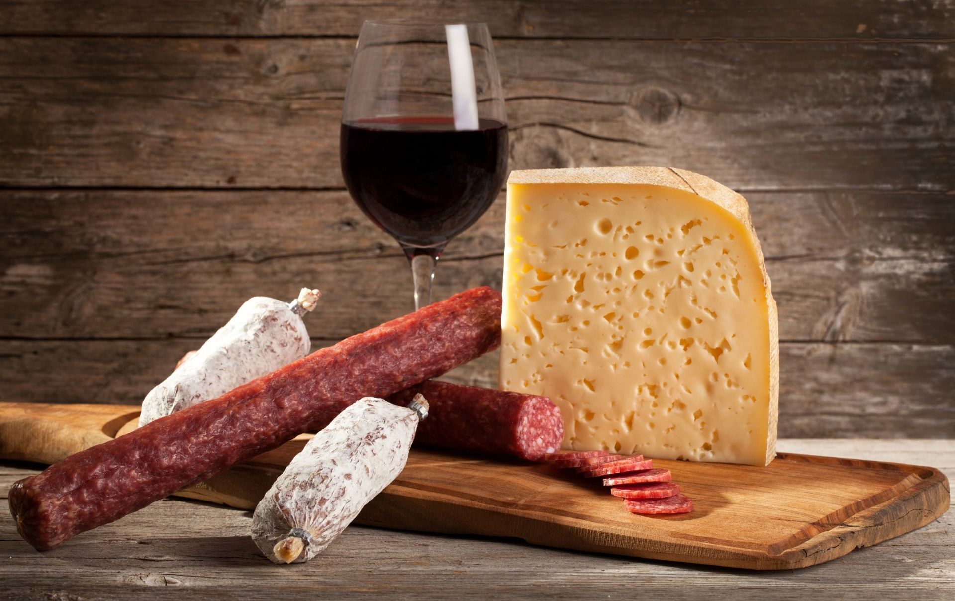 Une planche à découper en bois, sur laquelle on a disposé une grosse tranche de fromage, plusieurs tranches de salami et un verre de vin rouge.