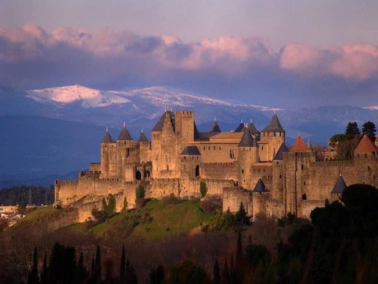 Un château historique doté de nombreuses tours et d’un mur d’enceinte, situé sur une colline, avec en toile de fond des montagnes et un ciel nuageux.