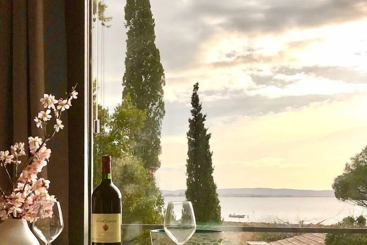 Uma mesa com uma garrafa de vinho e copos, um vaso com flores, e vista para o mar e árvores através de uma janela.