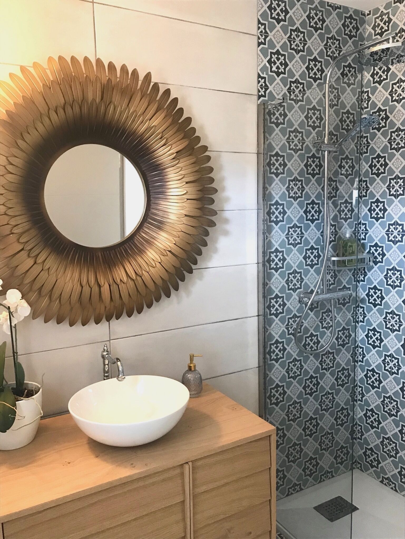Un baño con un espejo redondo, un lavabo blanco, un mueble de tocador de madera, una orquídea en maceta y una ducha con azulejos estampados.