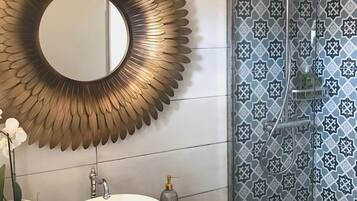 Un baño con un espejo redondo, un lavabo blanco, un mueble de tocador de madera, una orquídea en maceta y una ducha con azulejos estampados.