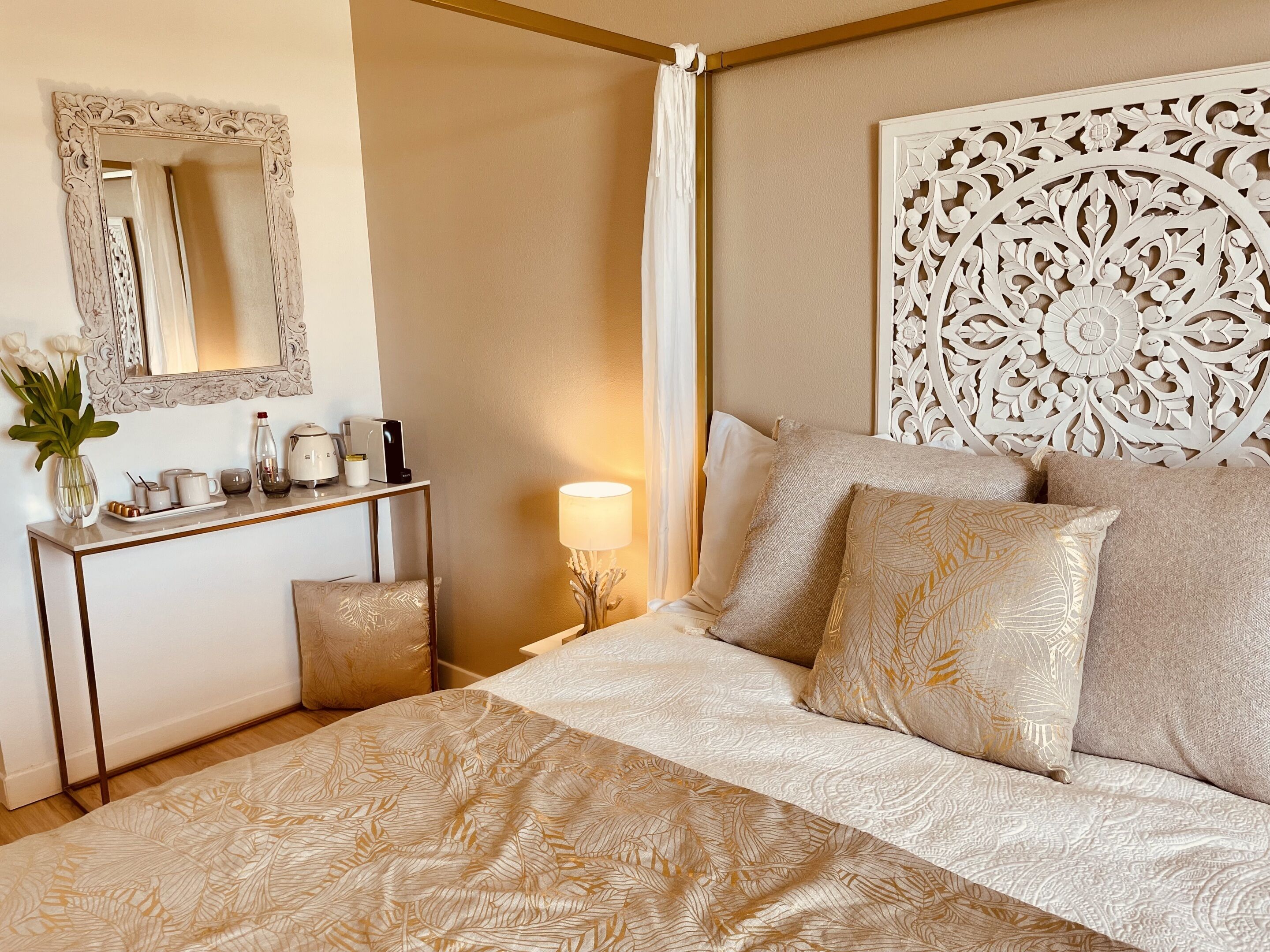 Een slaapkamer met een groot bed, een nachtkastje met een lamp, een spiegel en een decoratief hoofdeinde.