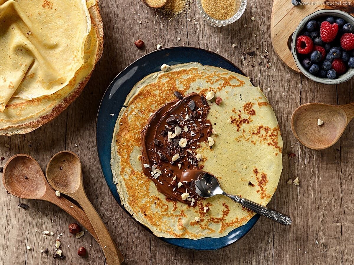 A plate of crepes with chocolate spread and nuts, accompanied by a bowl of berries and a wooden spoon.