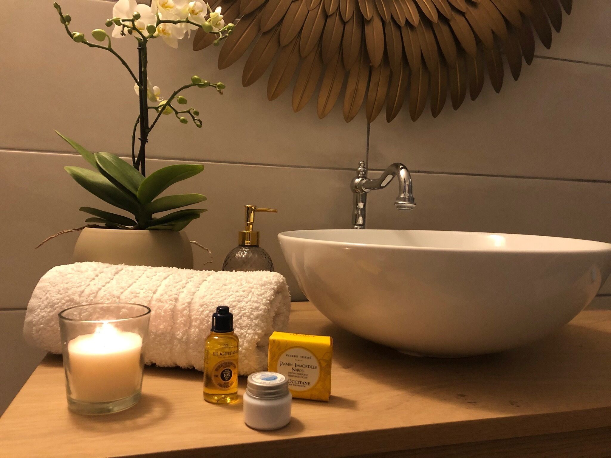 Un tocador de baño con un lavabo blanco, una planta en maceta, una toalla suave, una vela y artículos de aseo.