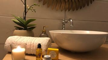 Un tocador de baño con un lavabo blanco, una planta en maceta, una toalla suave, una vela y artículos de aseo.