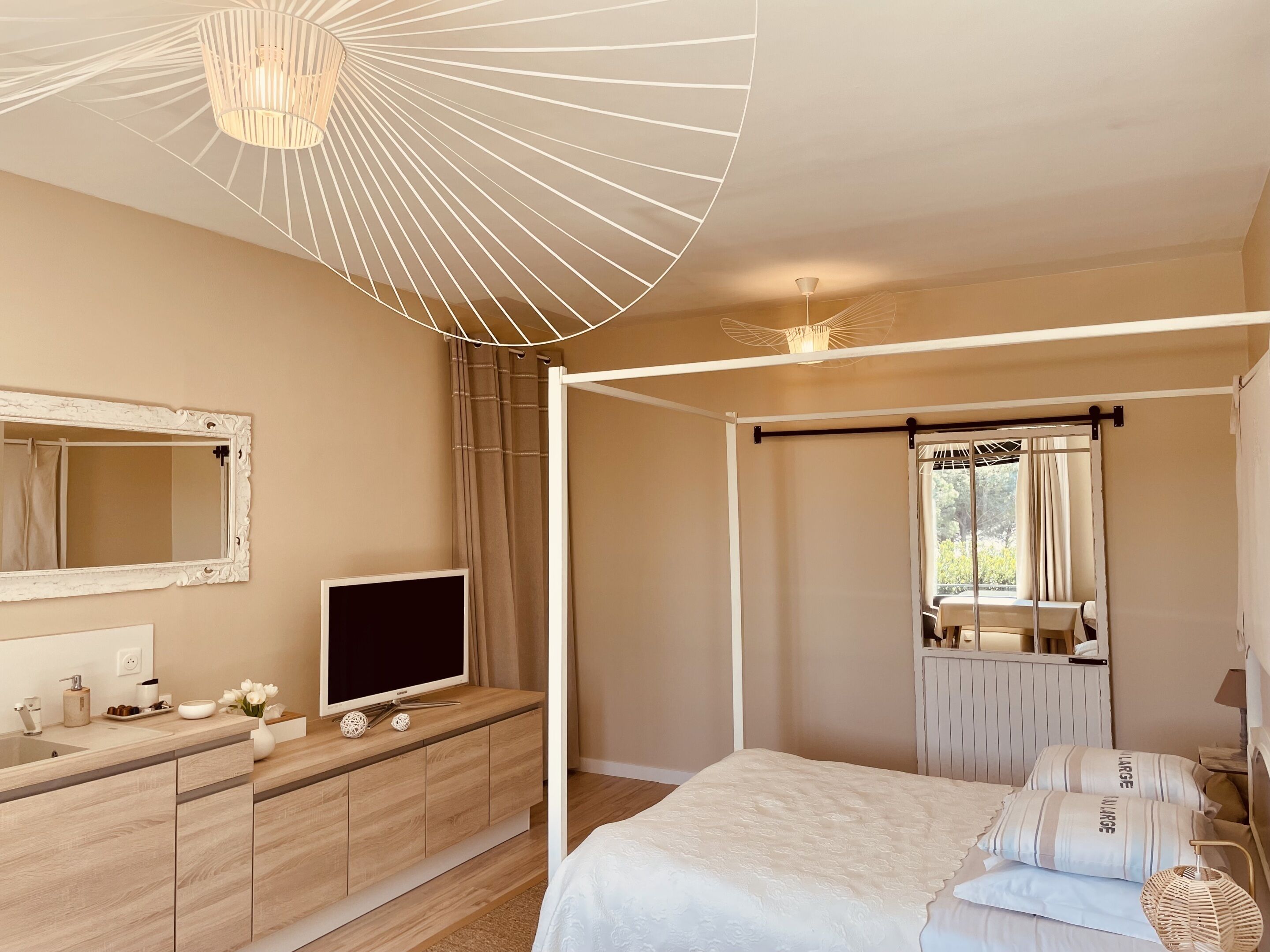 Une chambre à coucher avec un lit à baldaquin, un téléviseur posé sur une commode en bois, un grand miroir et une fenêtre avec des rideaux.