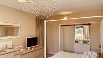 Un dormitorio con cama con dosel, un televisor sobre una cómoda de madera, un espejo grande y una ventana con cortinas.