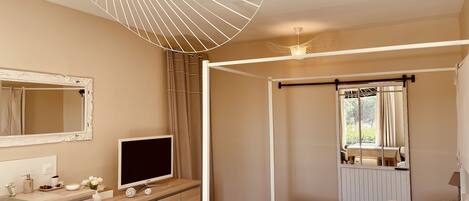 Une chambre à coucher avec un lit à baldaquin, un téléviseur posé sur une commode en bois, un grand miroir et une fenêtre avec des rideaux.