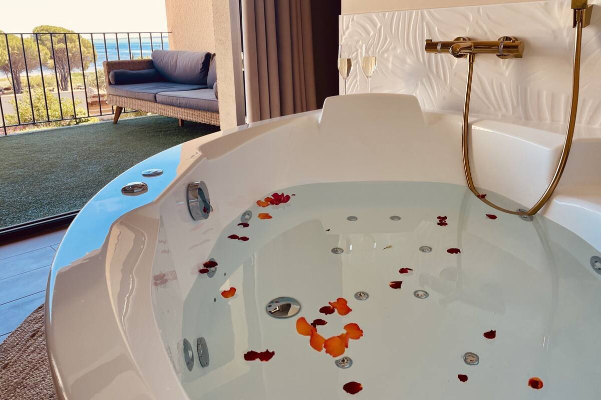Uma banheira grande e branca com pétalas de rosa, um chuveiro dourado e vista para o mar através de uma varanda.