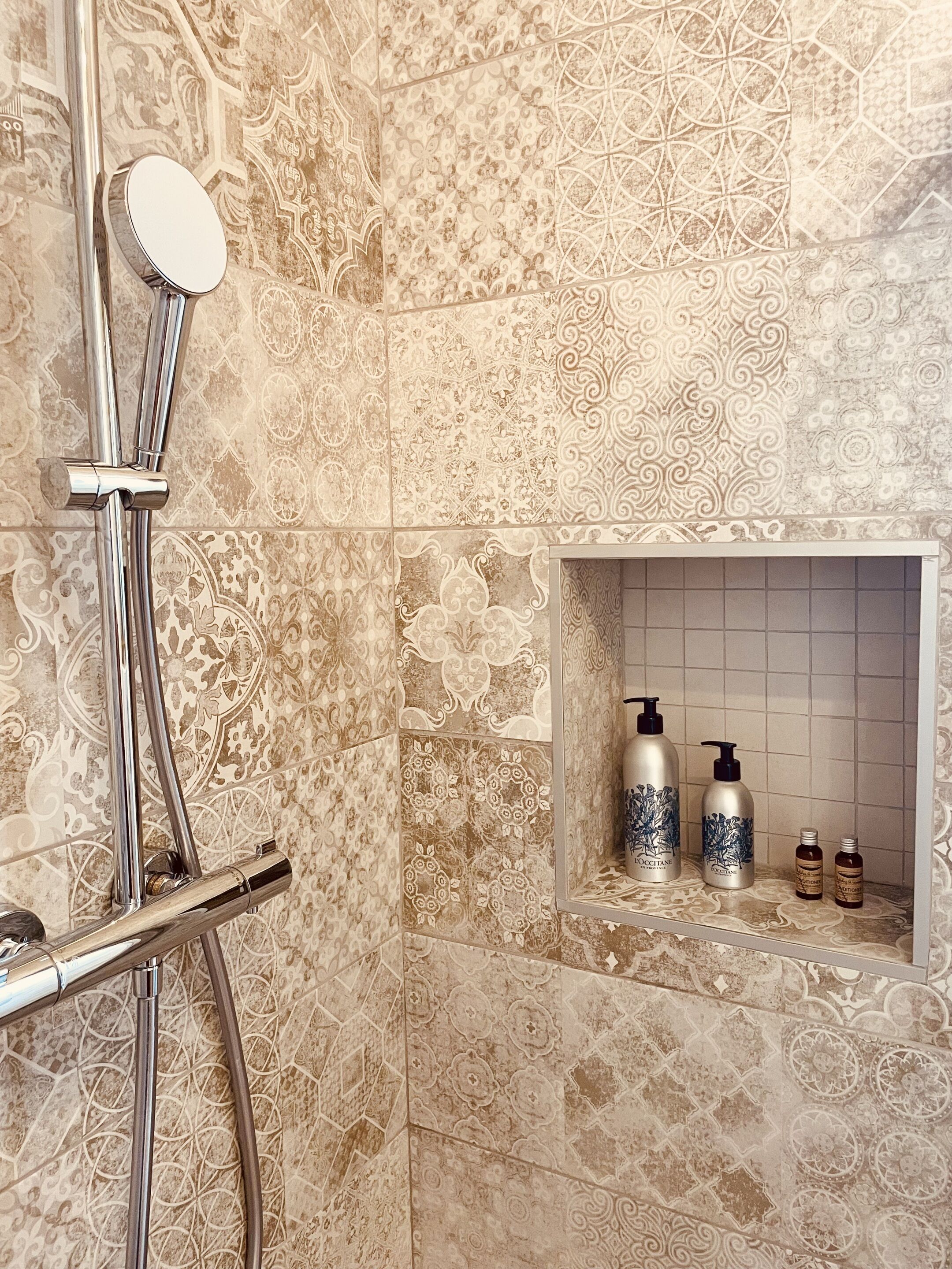 A bathroom with ornate tile patterns, a chrome showerhead, and a shelf with toiletries.