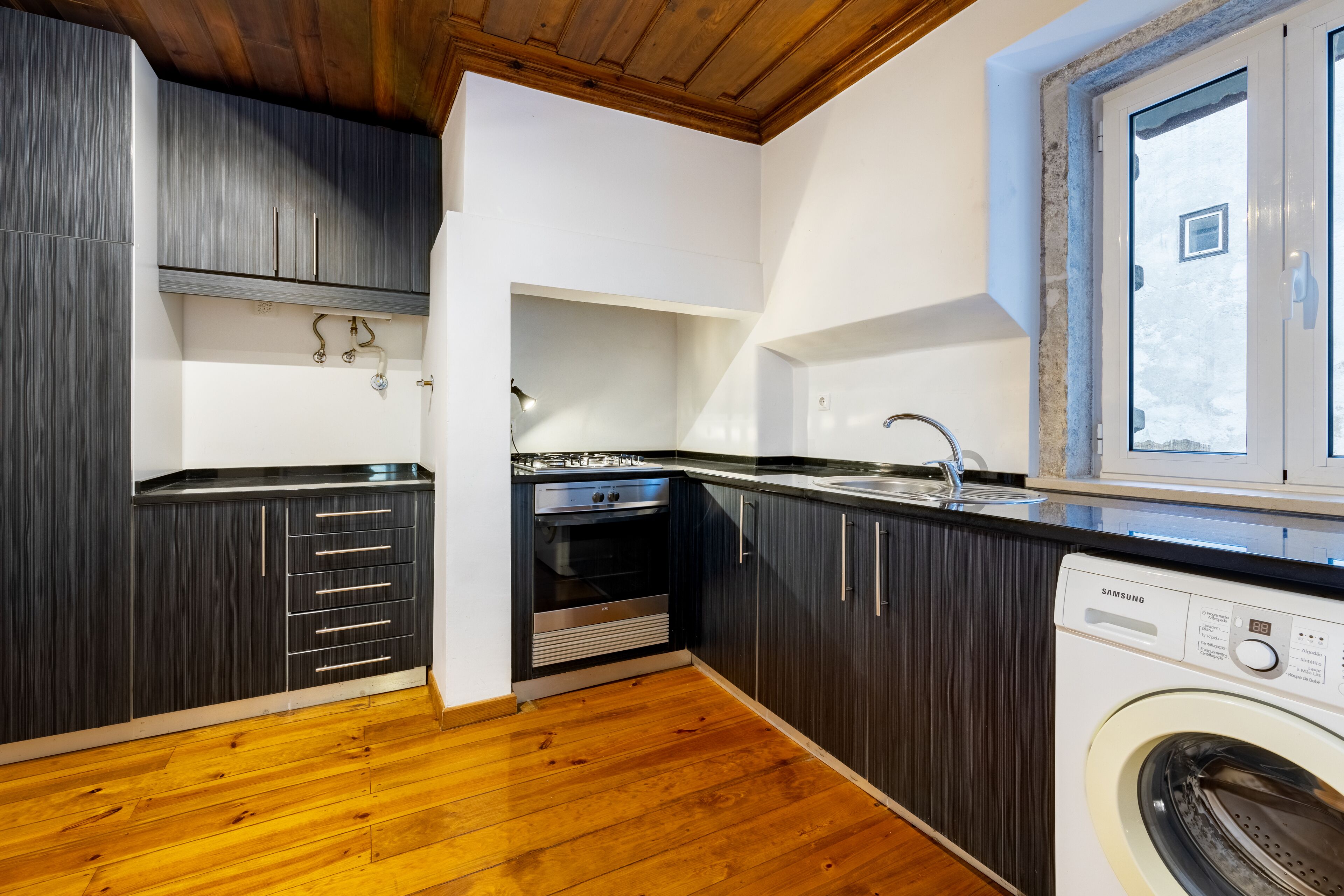 A modern kitchen with dark cabinetry, a built-in oven, a washing machine, and a window with a view.