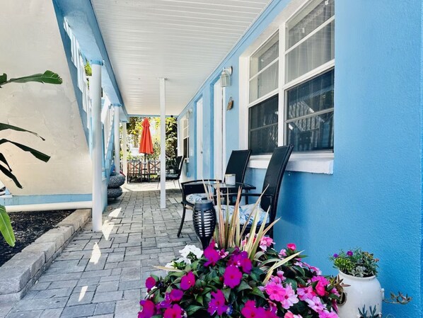 A covered patio area with a blue exterior wall, a brick floor, and outdoor furniture including two chairs and a table. - Blue Water Beachside Rental 7 (Indian Rocks Beach)