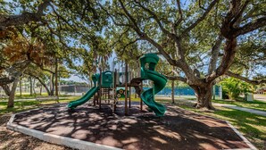 A playground with a slide and climbing structures, surrounded by trees and a paved path. - Blue Water Beachside Rental 7 (Indian Rocks Beach)