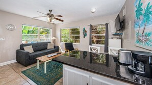 A compact living space with a kitchenette, a ceiling fan, a dining area, and large windows offering a view of greenery. - Blue Water Beachside Rental 7 (Indian Rocks Beach)