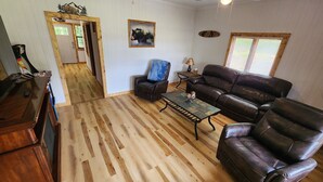 A living room with a wooden floor, a leather sofa, a coffee table, and a chair. There is a door leading to another room. - Daisy's Riverside Log Cabin with 2 kayaks, Arcade, hot tub & recliners. (Pigeon Forge)