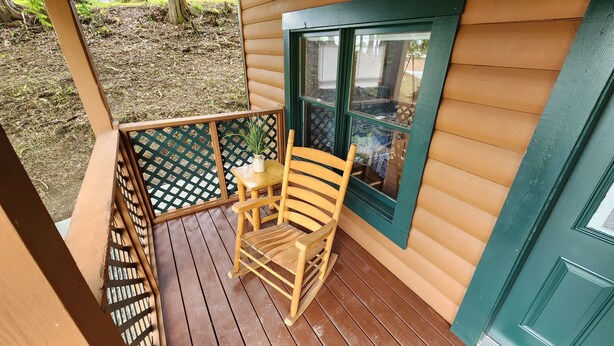 A wooden deck with a rocking chair and a table, a window with a view of a house, and a green lattice fence. - Daisy's Riverside Log Cabin with 2 kayaks, Arcade, hot tub & recliners. (Pigeon Forge)