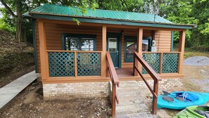 A small cabin with a green roof, a porch with a wooden railing, and a brick foundation. - Daisy's Riverside Log Cabin with 2 kayaks, Arcade, hot tub & recliners. (Pigeon Forge)