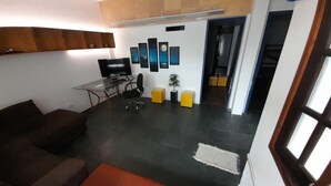 Un salón con una mesa de vidrio, una silla de oficina negra y un televisor. Hay dos sofás y un cojín naranja pequeño. El cuarto tiene piso de baldosas y una pared con cuadros enmarcados.