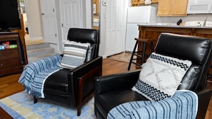 Una sala de estar con dos sillones de cuero negro, un mueble de madera, un televisor y una zona de cocina con alacenas de madera y un microondas.