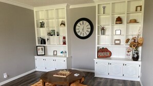 A living room with a wooden coffee table, cushioned ottomans, white shelving units, and a clock on the wall. - Country Retreat north of Chandler (Tryon)