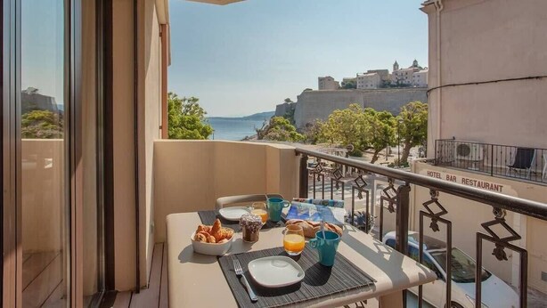 A balcony with breakfast setup overlooking a coastal view, including a hotel bar restaurant in the background. - Casa Aliméa (Calenzana)