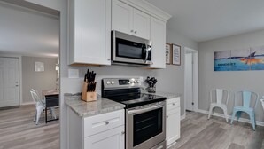 Eine moderne Küche mit weißen Schränken, einer eingebauten Mikrowelle und einem Edelstahlbackofen. Im Essbereich stehen zwei Stühle und es gibt eine Kücheninsel mit einem Messerblock.