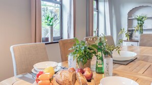 A dining table with a variety of food items, including bread, fruits, and a bottle of green juice. There are chairs around the table, and a window with plants visible in the background. - 4 bedroom accommodation in Llysfaen, near Colwyn Bay (Llysfaen, near Colwyn Bay)