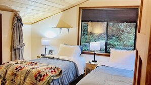 Quarto com duas camas, janela com persianas, ventilador de teto e abajur em uma mesa de cabeceira.