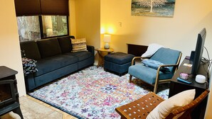 Uma sala de estar acolhedora com um sofá azul escuro, um tapete estampado, uma poltrona azul, uma mesinha de madeira com uma luminária e um quadro de uma árvore na parede.