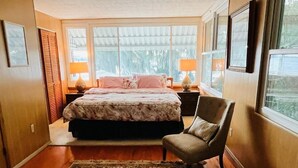 Un dormitorio con una cama grande, dos lámparas de noche, una silla y vistas a zonas verdes a través de las ventanas.